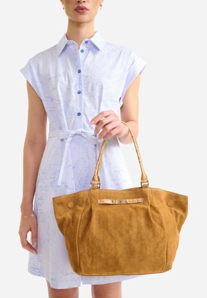 Donna che indossa un abito azzurro a righe sottili, che tiene in mano una grande borsa tote in suede color cammello con doppi manici e dettaglio con cinturino anteriore.