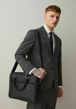 Bărbat în costum gri și cravată în dungi, ținând o servietă neagră din piele, arătând un tatuaj floral detaliat pe mână, pe fundalul unui perete simplu deschis la culoare.