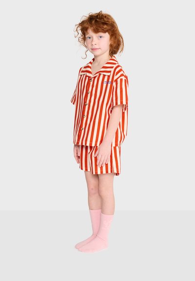 Enfant aux cheveux roux bouclés debout portant un t-shirt à manches courtes rayé orange et blanc et un short assorti avec des chaussettes roses sur fond clair.