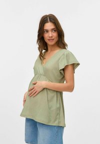 Top premaman verde chiaro con maniche corte a volant, design avvolgente, tessuto testurizzato e aderente sotto il seno; abbinato a pantaloni corti in denim blu.