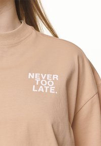 Béžová mikina s kulatým výstřihem. Obsahuje bílý nápis "NIKDY NENÍ POZDĚ." Tkanina vypadá hladce a má mírně volnější střih.