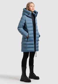 Sininen toppatakki, jossa on ylisuurikokoinen huppu, tikattu muotoilu, vetoketjulliset taskut ja suora leikkaus, yhdistetty mustiin leggingseihin ja nauhallisiin saappaisiin.