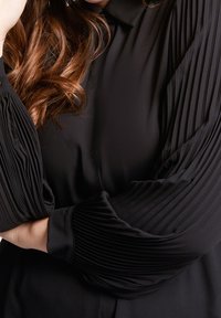 Lång svart blus med plisserade ärmar. Slät tyg med en strukturerad krage och knappstängning, med mjuka, flödande draperingar.