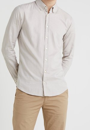 Muž v béžové košili na knoflíky s límečkem a hnědých kalhotách stojí se založenýma rukama za zády na bílém pozadí.