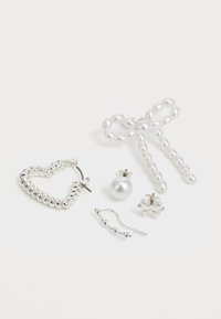 Pięć elementów biżuterii: srebrna kolczyk w kształcie serca, perłowe kolczyki, kolczyki w kształcie kokardy z pereł, mały srebrny kwiatek oraz falisty srebrny kolczyk, wszystkie o różnych teksturach.