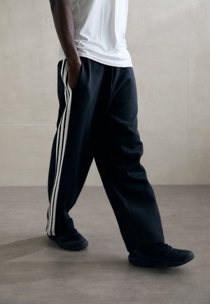 Persona con pantalones deportivos negros con rayas blancas, zapatillas negras y camiseta blanca, de pie frente a un fondo gris liso.