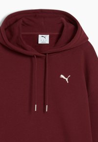 Burgundy hætte sweatshirt i blødt stof har en kangaroo lomme og et lille hvidt broderet logo på brystet. Sølv snørebånd hænger ned.