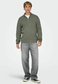 Olijfgroene sweatshirt met halve ritssluiting, gecombineerd met lichtgrijze spijkerbroek en zwarte sneakers, staand op een neutrale achtergrond.