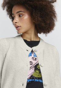 Cárdigan color crema con tejido texturizado, cuello redondeado y dos botones en el cierre. Lleva puesto un camiseta negra gráfica con un diseño colorido.