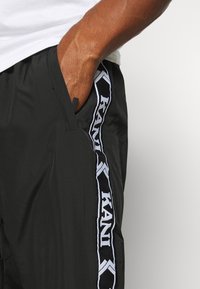 Hand van een persoon in de zak van zwart sportshorts met een zwart-witte "KANI" merkstreep aan de zijkant, draag een wit shirt.