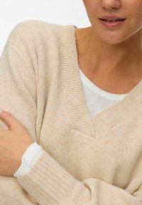 Beżowy sweter z dekoltem w serek, z prążkowanymi mankietami i dołem, wykonany z miękkiej mieszanki wełny, nałożony na biały, strukturalny, długi rękaw koszulę.