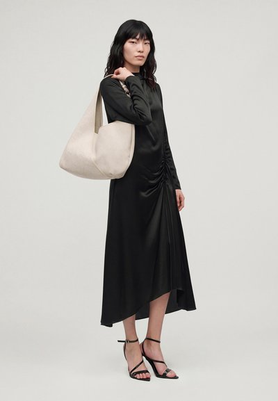 Un mannequin portant une robe midi noire et des chaussures à talons à brides porte un grand sac à bandoulière couleur crème devant un fond gris uni.