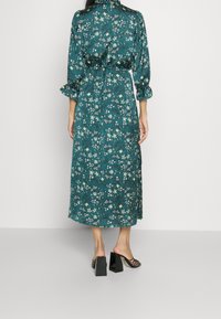 Vestido midi de flores en color teal con mangas largas, tejido recogido en la cintura y sandalias de tacón negras. El vestido presenta un patrón de flores en blanco y amarillo.