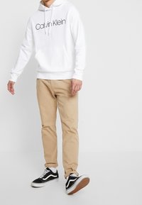 Sudadera blanca con capucha y logotipo negro de "Calvin Klein", pantalones beige y zapatillas Vans negras con detalles en blanco y suelas texturizadas.