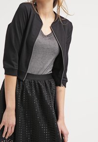 Czarna kurtka bomberka z srebrnym zamkiem, lekka tkanina i rękawy do trzech kwartałów na szarym T-shircie, w połączeniu z teksturowaną czarną spódnicą.