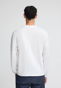 Vit långärmad skjorta i bomull med rund halsringning och avslappnad passform. Bärs med mörkblå byxor, visar bakifrån.