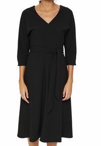 Robe midi noire avec col en V, manches trois-quarts, et ceinture en tissu nouée à la taille, portée par une personne debout contre un fond blanc.