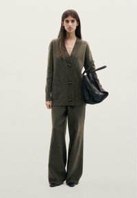 Cardigan vert olive surdimensionné avec un col côtelé texturé, une fermeture à boutons sur le devant et deux grandes poches, associé à un pantalon large olive et un sac noir.