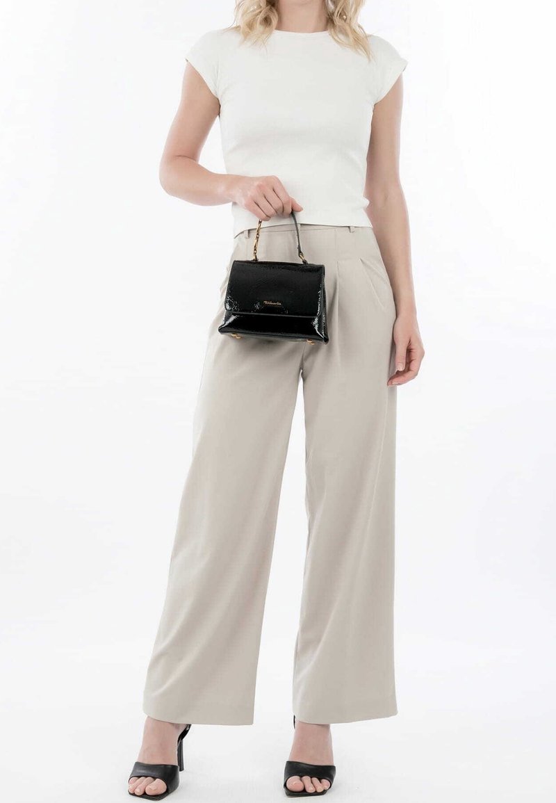 Femme portant un haut blanc à manches courtes, un pantalon beige à jambes larges, des sandales noires à talons et tenant un petit sac à main noir par la poignée.
