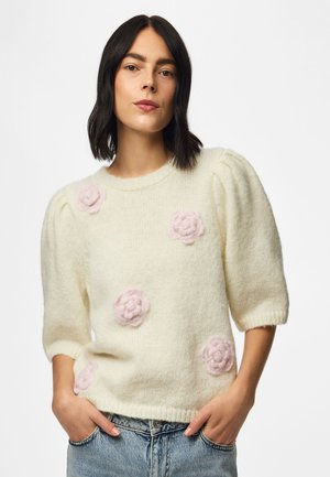 Femme aux cheveux foncés portant un pull en maille crème avec des manches bouffantes et des appliques florales roses, associé à un jean bleu clair.
