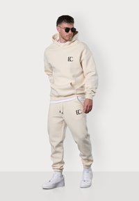 Beige Kapuzenpullover und passende Jogginghose, beide aus weichem Stoff gefertigt. Mit schwarzem Logo auf der Brust und dem Bein, elastischen Bündchen und Kordelzug.