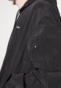 Veste légère noire avec une texture froissée, dotée d'une capuche, d'une fermeture éclair à l'avant et d'un discret logo sur la poitrine.
