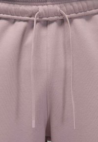 Lähikuva liilanväristen collegehousujen vyötärönauhasta, jonka mukana tulevat kiristysnauhat roikkuvat löysästi kangaspinnan edessä.