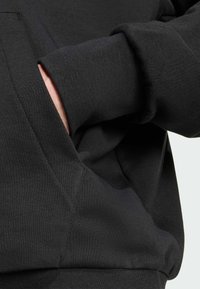 Sudadera negra con un tejido texturizado, que presenta un bolsillo frontal y puños acanalados, exhibiendo un diseño simple y funcional.