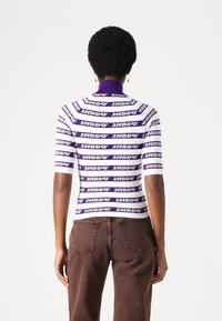 Top aderente a maniche corte di colore bianco con strisce orizzontali viola, con testo "2NDAY". Collo a coste e texture morbida, abbinato a pantaloni marroni.