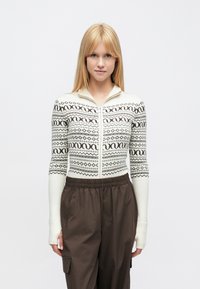 Gaiši krēmkrāsas džemperis ar garām piedurknēm un brūniem ģeometriskiem rakstiem, ar rāvējslēdzēju priekšpusē un ribotām aprocēm, kopā ar tumši brūnām biksēm.