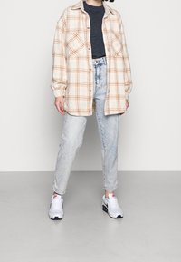 Chemise en flanelle à carreaux beige et orange, portée sur un pull gris foncé, associée à un jean bleu clair et des baskets blanches. Texture lisse, coupe décontractée.