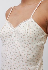 Camiseta sin mangas floral con ribete de encaje. Tejido blanco roto con pequeños patrones de flores rosas y verdes. Tirantes ajustables tipo espagueti. Textura suave.