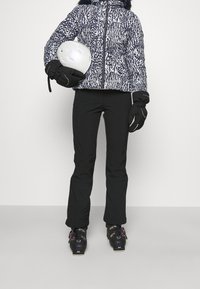 Tenue de ski : doudoune à motifs avec capuche, pantalon noir, chaussures de ski noires avec accents roses, gants et casque blanc.