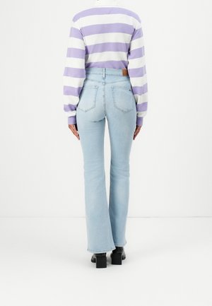 Personne portant un jean évasé bleu clair, des bottes noires à talons et une chemise à manches longues rayée lavande et blanche, debout devant un fond blanc.