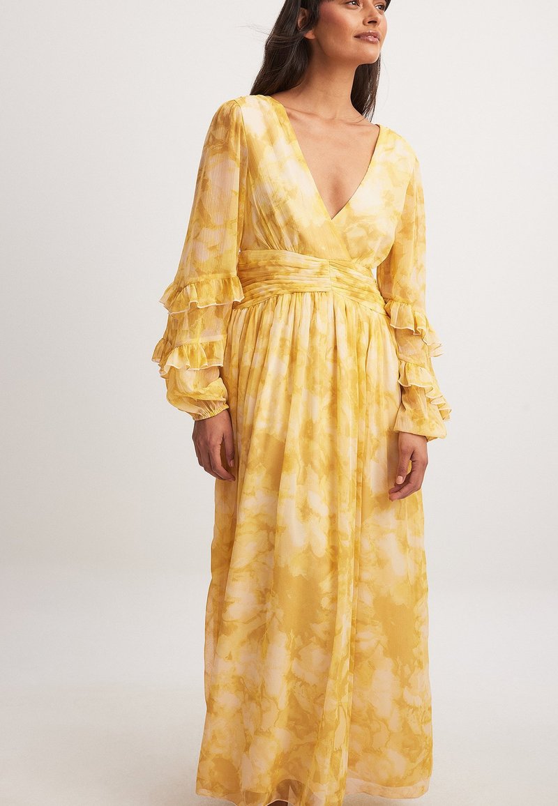 Keltainen kukkakuosis maxi-mekko, jossa on V-pääntie, pitkät röyhelöillä varustetut hihat ja vyöllinen vyötärö. Kevyt kangas, jossa on virtaava ja tekstuuriltaan mielenkiintoinen ilme.