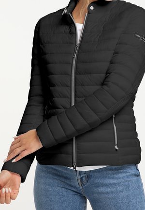 Mujer que lleva una chaqueta acolchada negra con cremallera frontal y bolsillos, ajustándose la manga mientras lleva vaqueros azules y una camisa blanca debajo.