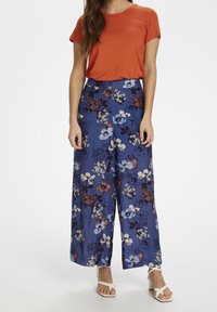 Oranje t-shirt met korte mouwen, gecombineerd met een lichtblauwe broek met wijde pijpen en een bloempatroon van rode en witte bloemen. Witte sandalen met hakken en banden.