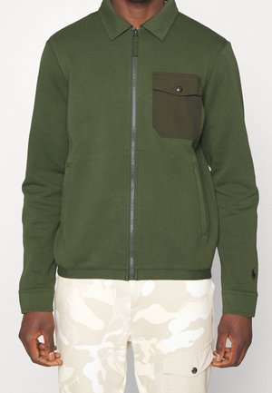 Mand iført olivengrøn lynlåstrøje med mørkegrøn brystlomme med knapper og beige camouflage cargobukser.