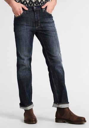 Homme portant un jean denim bleu foncé avec les revers retroussés, des bottines en cuir marron et les mains dans les poches, sur un fond uni.