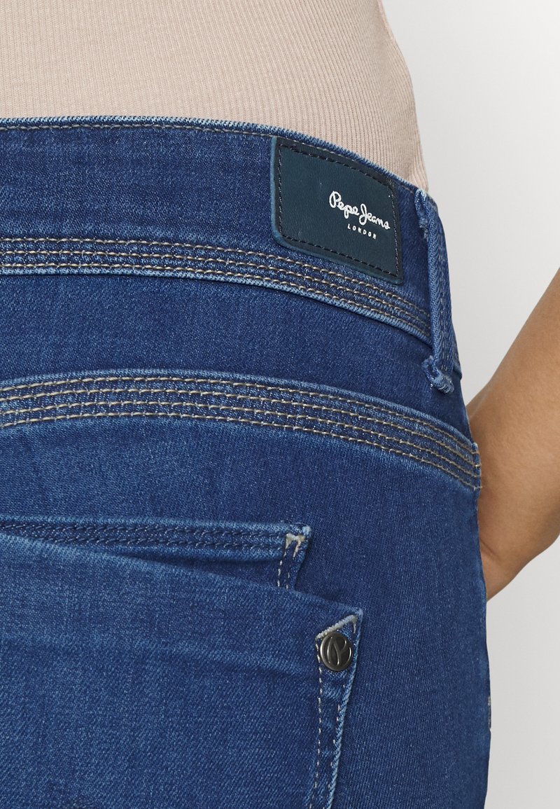 Gros plan sur l'arrière d'un jean en denim bleu avec double piqûre, étiquette noire Pepe Jeans London, et une partie du bras d'une personne portant un haut beige.