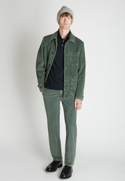 Chaqueta de pana verde con bolsillos frontales, combinada con pantalones a juego y una camiseta de polo negra. Un gorro de punto gris y zapatos negros completan el look.