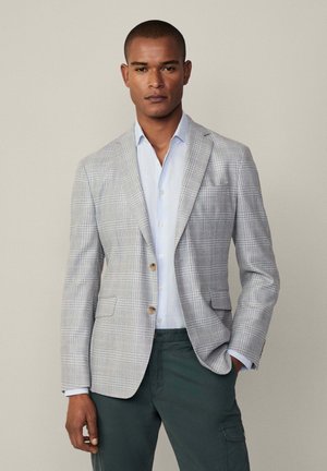Blazer à carreaux gris en tissu texturé, porté sur une chemise bleu clair. Le blazer a deux boutons et des poches latérales.