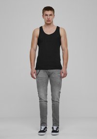 Czarny bezrękawnik, szare zniszczone dżinsy i czarne sneakersy z białymi podeszwami. Model stoi na neutralnym tle.