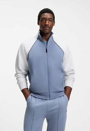 Man draagt een lichtblauw jack met ritssluiting en witte mouwen, gecombineerd met een lichtblauwe broek, staand met handen in de zakken.