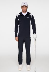 Marinblå zip-up sportstopp med vita sidodetaljer, kombinerad med marinblå byxor och vita skor. Bär en vit keps och håller en golfklubba.