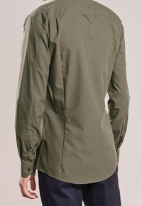 Camisa de manga larga color verde oliva, tela lisa, diseño ajustado, cuello puntiagudo, puños con botones, dobladillo redondeado, detalle minimalista en la parte trasera.