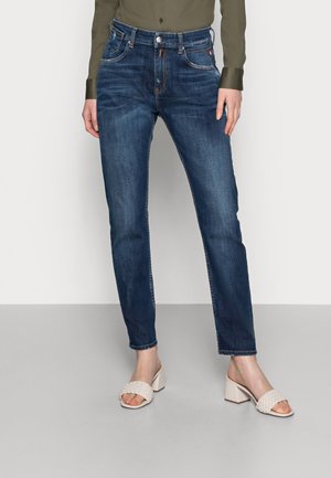 Frau trägt dunkelblaue Slim-Fit-Jeans, olivgrünes langärmeliges Shirt und weiße, blockabsatzige, geflochtene Sandalen auf grauem Boden.