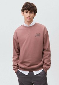 Felpa rosa con colletto a coste, caratterizzata da una piccola grafica stampata sul petto, indossata sopra una camicia bianca con colletto.