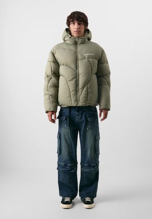 Chaqueta acolchada verde oliva con cuello alto, cremallera frontal y costuras geométricas, combinada con pantalones cargo azules y zapatillas negras.