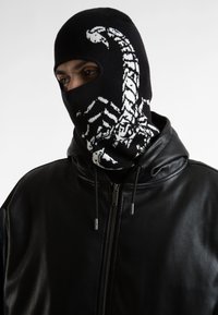 Balaclava nera in maglia con un design di scorpione bianco, indossata con una giacca in pelle nera con zip. Tessuto elastico e vestibilità aderente.